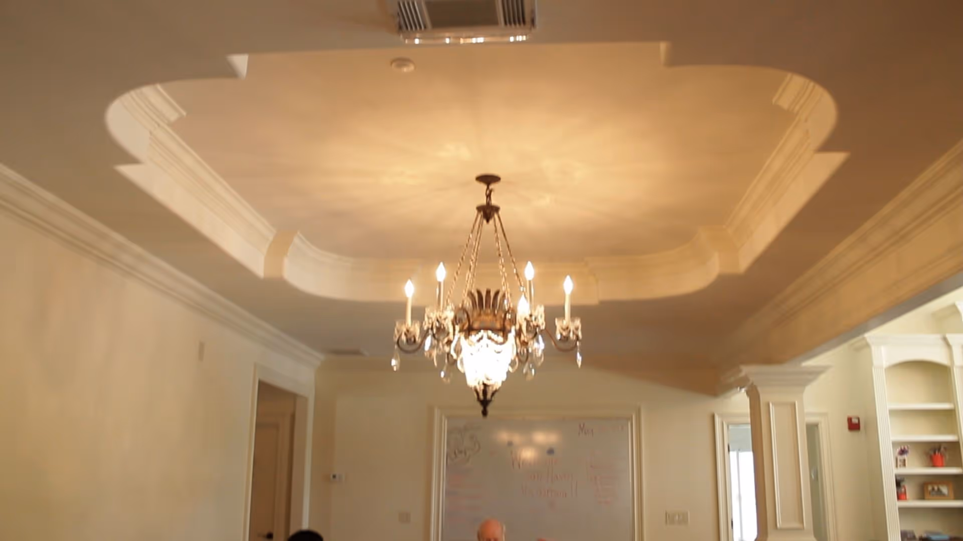 An elegant chandelier hanging from a decorative recessed ceiling in a room with cream-colored walls. Below the chandelier, a whiteboard with writing is visible on the wall, and part of a person’s head is seen at the bottom of the image. The room has white trim and built-in shelves on the right side.