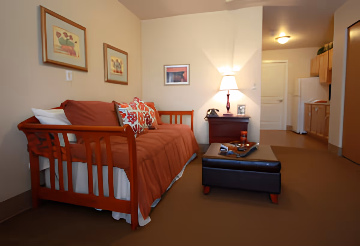 A cozy living area in an assisted living facility featuring a wooden daybed with red cushions and patterned pillows, a small wooden side table with a lamp and a vintage telephone, a dark ottoman coffee table with decorative items, and a kitchenette with wooden cabinets and a white refrigerator in the background. Three framed artworks hang on the beige walls.