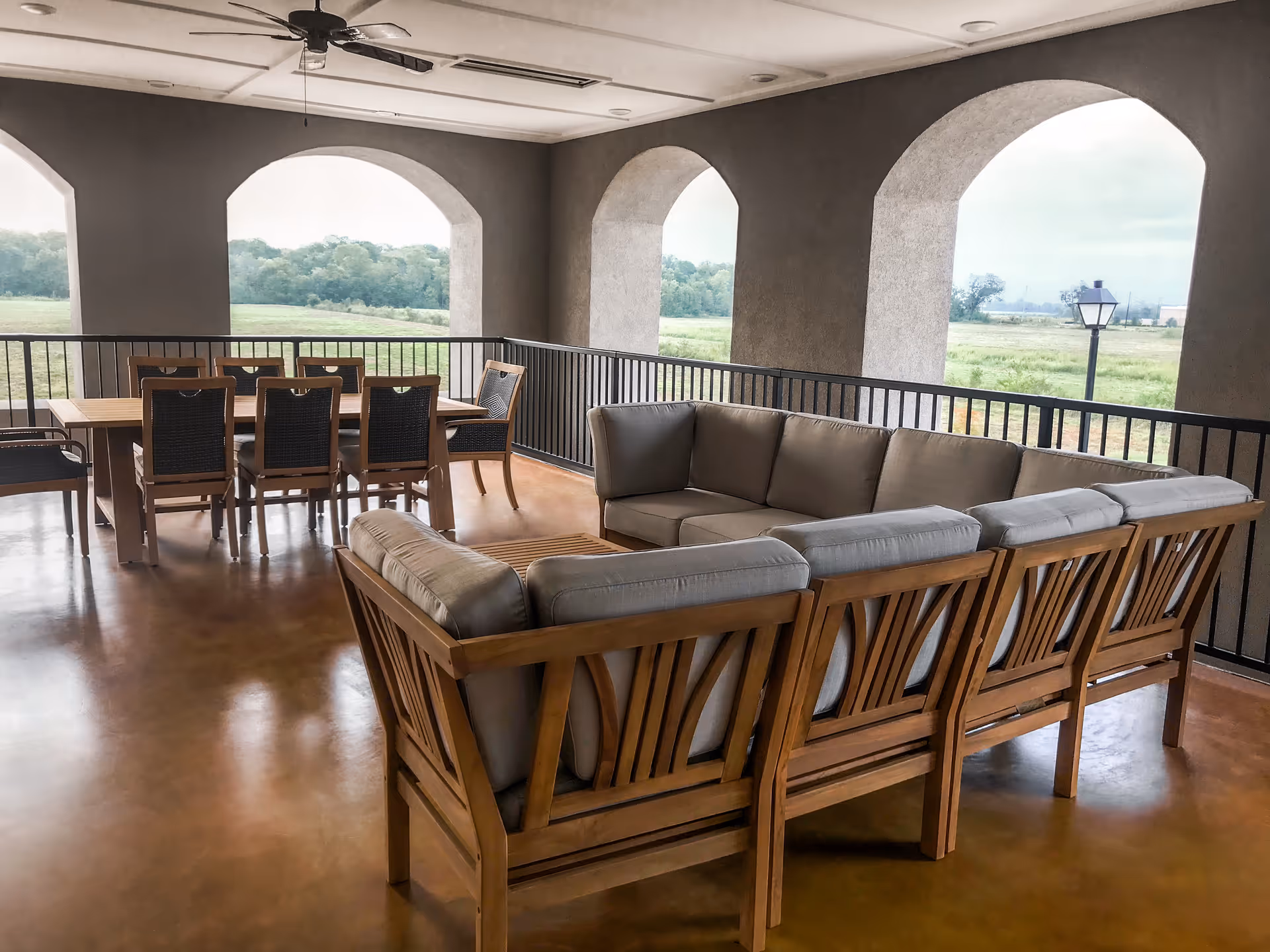 Covered outdoor patio area with a wooden sectional sofa with gray cushions and a wooden dining table with six chairs. The patio has arched openings overlooking a green field and trees in the distance. A ceiling fan is mounted above.