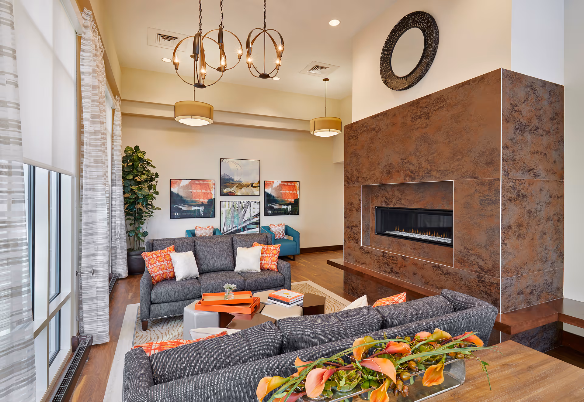 A modern living room with two gray sofas adorned with orange and white pillows, a rectangular coffee table with orange trays and books, a large brown textured fireplace with a circular mirror above it, three abstract paintings on the wall, a tall potted plant in the corner, and large windows with sheer curtains letting in natural light.