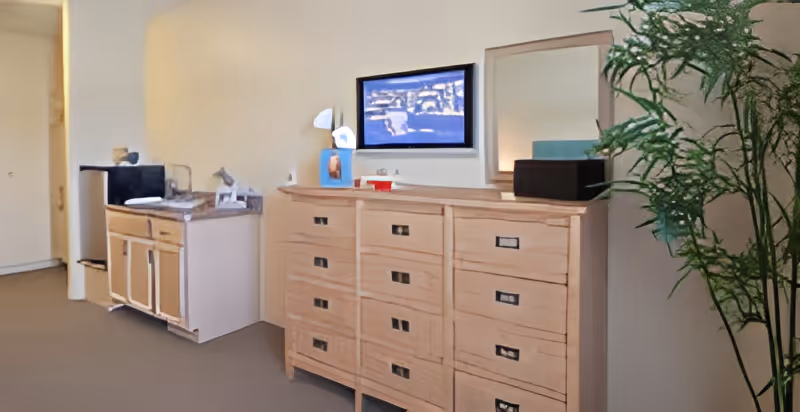 Interior view of a room in Danbury Columbus facility showing a wooden dresser with multiple drawers, a mirror above it, a small kitchenette area with a microwave and mini fridge, and a tall green plant on the right side.