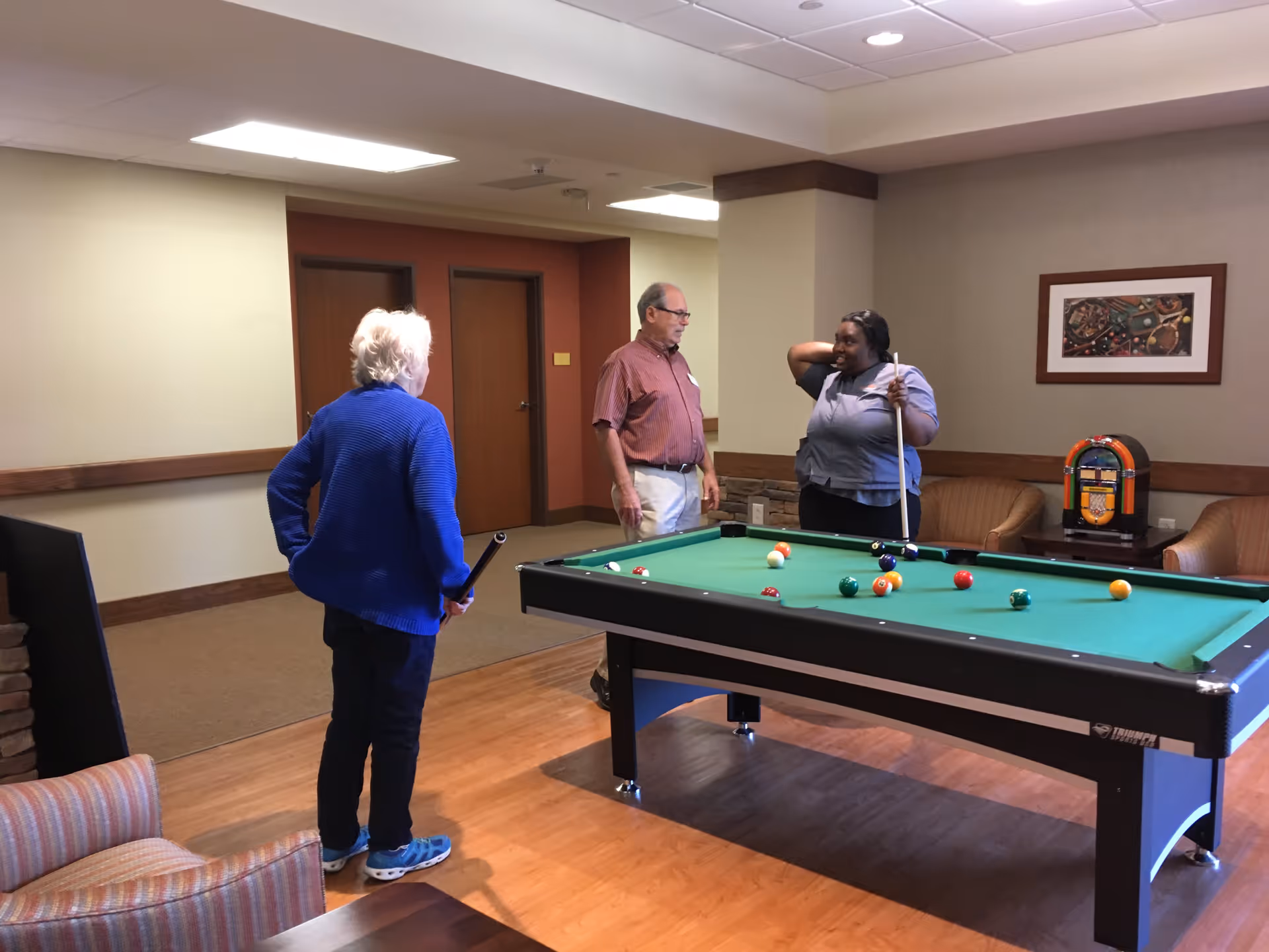 Three people standing around a pool table in a common room. One person is holding a pool cue and appears to be playing, while the other two watch and talk. The room has wooden flooring, beige walls, a framed picture, a jukebox, and comfortable chairs.