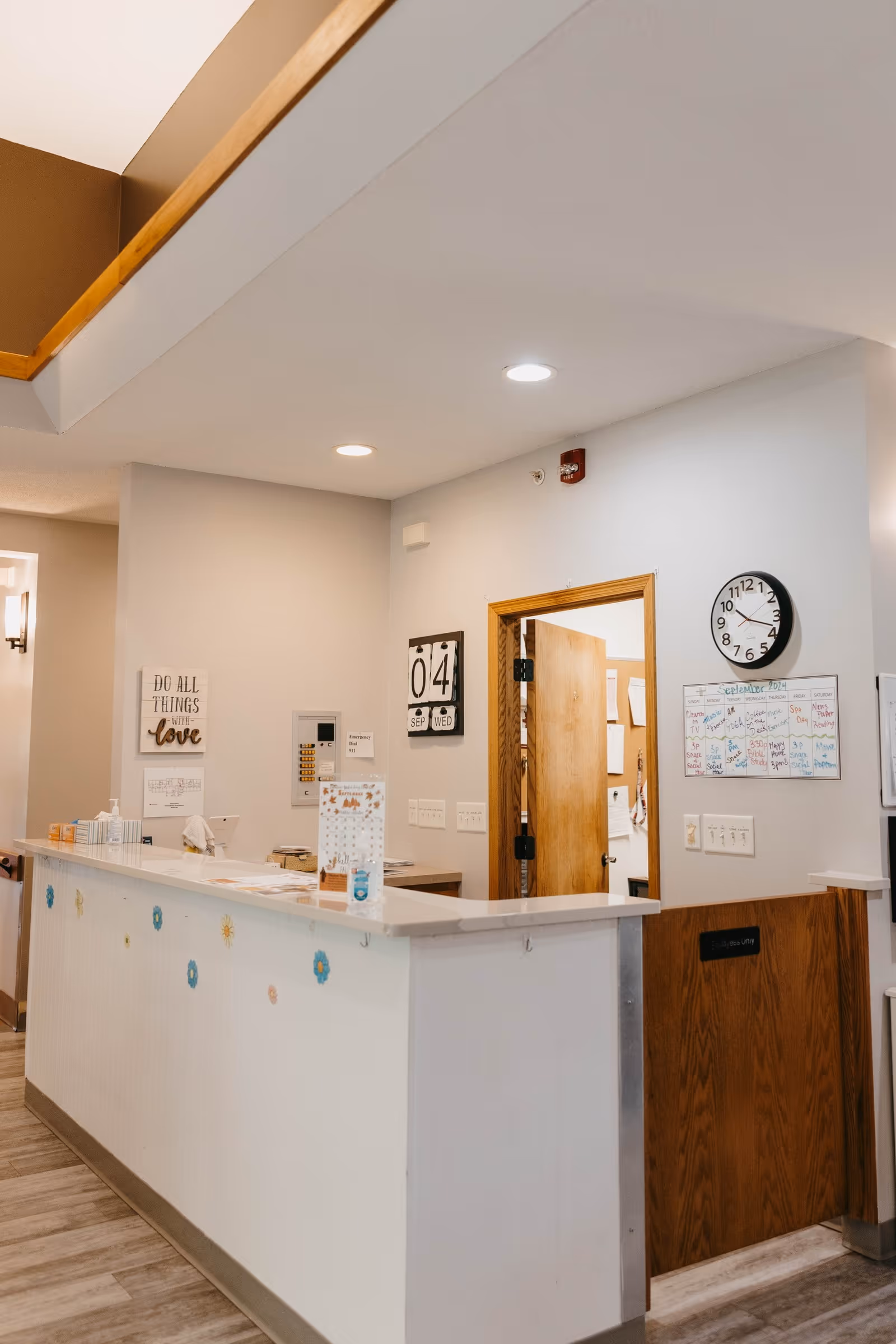 Reception area of an assisted living facility with a white counter decorated with small flower stickers. Behind the counter is a wooden door slightly open, a wall clock showing 11:10, a calendar, and a sign that reads 'Do all things with love'. The walls are light-colored and the floor has wood-like tiles.