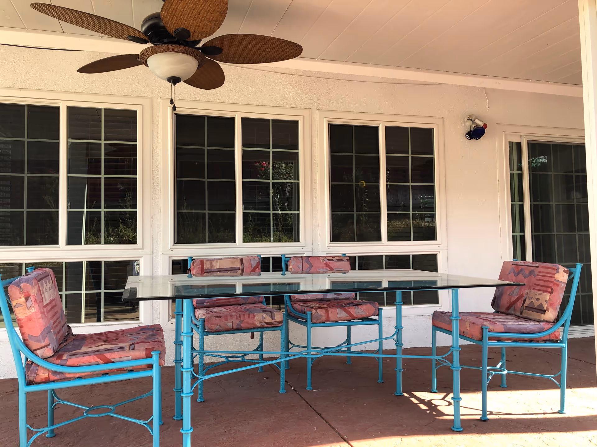 Outdoor patio area with a glass-top table and four blue metal chairs with patterned cushions. The patio is covered with a ceiling that has a ceiling fan with brown blades. Behind the table are large windows and a sliding glass door.