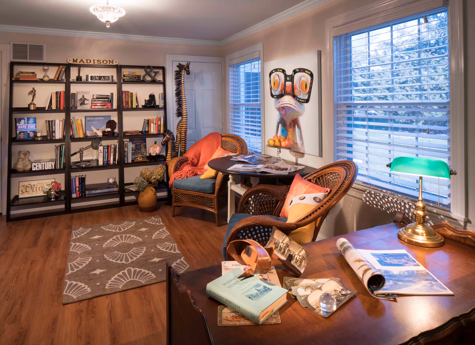 Cozy living room-style sitting area with bookshelves, wicker chairs around a small table, a desk with a green banker’s lamp, and large windows with blinds.