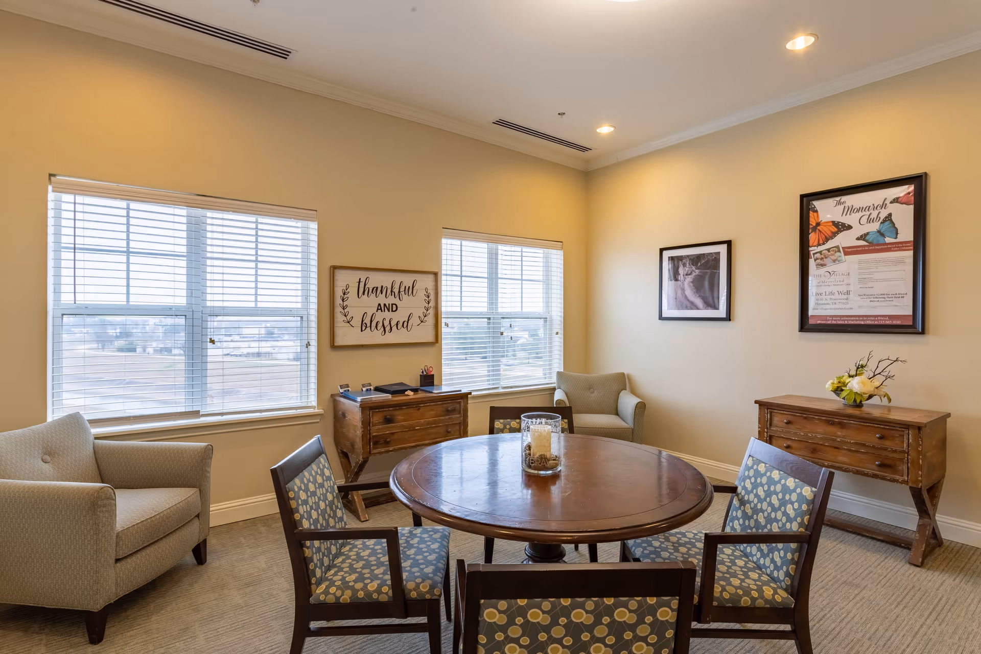 A cozy sitting room with a round wooden table surrounded by four patterned chairs. Two beige armchairs are placed near two large windows with white blinds. The walls are painted light yellow and decorated with framed pictures and a sign that reads 'thankful AND blessed.' Two wooden side tables hold small decorative items and office supplies.