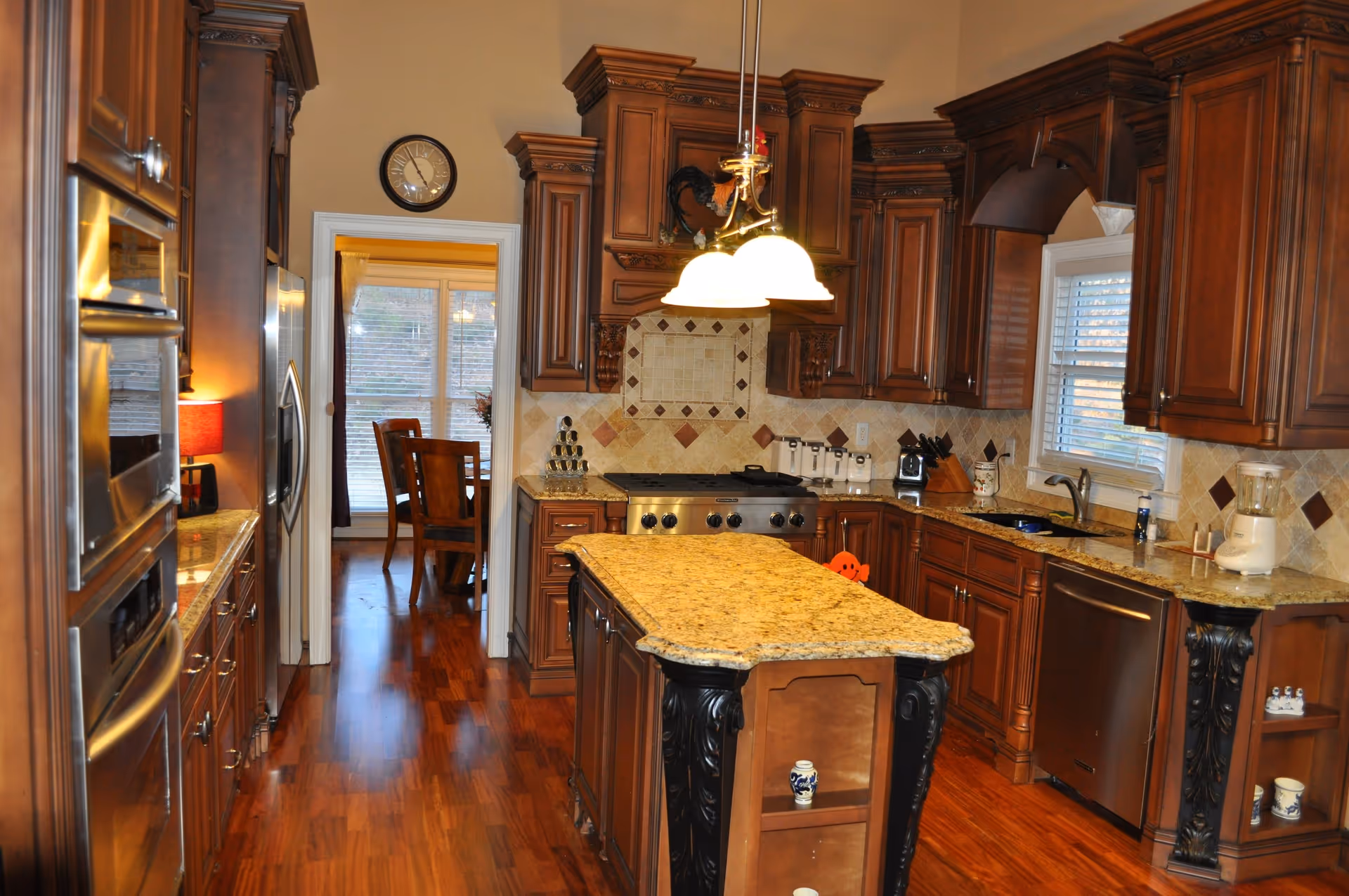 A spacious kitchen with wooden cabinets, granite countertops, and a central island. The kitchen features stainless steel appliances including a refrigerator, oven, and dishwasher. There is a window above the sink with white blinds, and a hanging light fixture above the island. In the background, a dining area with wooden chairs and a large window with blinds is visible.