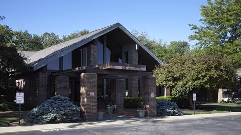 Exterior view of a brick building with a peaked roof and large windows, surrounded by trees and shrubs under a clear blue sky.