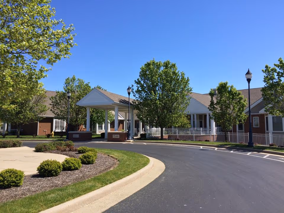 Exterior view of a senior living facility with a curved driveway, landscaped bushes, trees, and a covered entrance. The building has a brick and white facade under a clear blue sky.