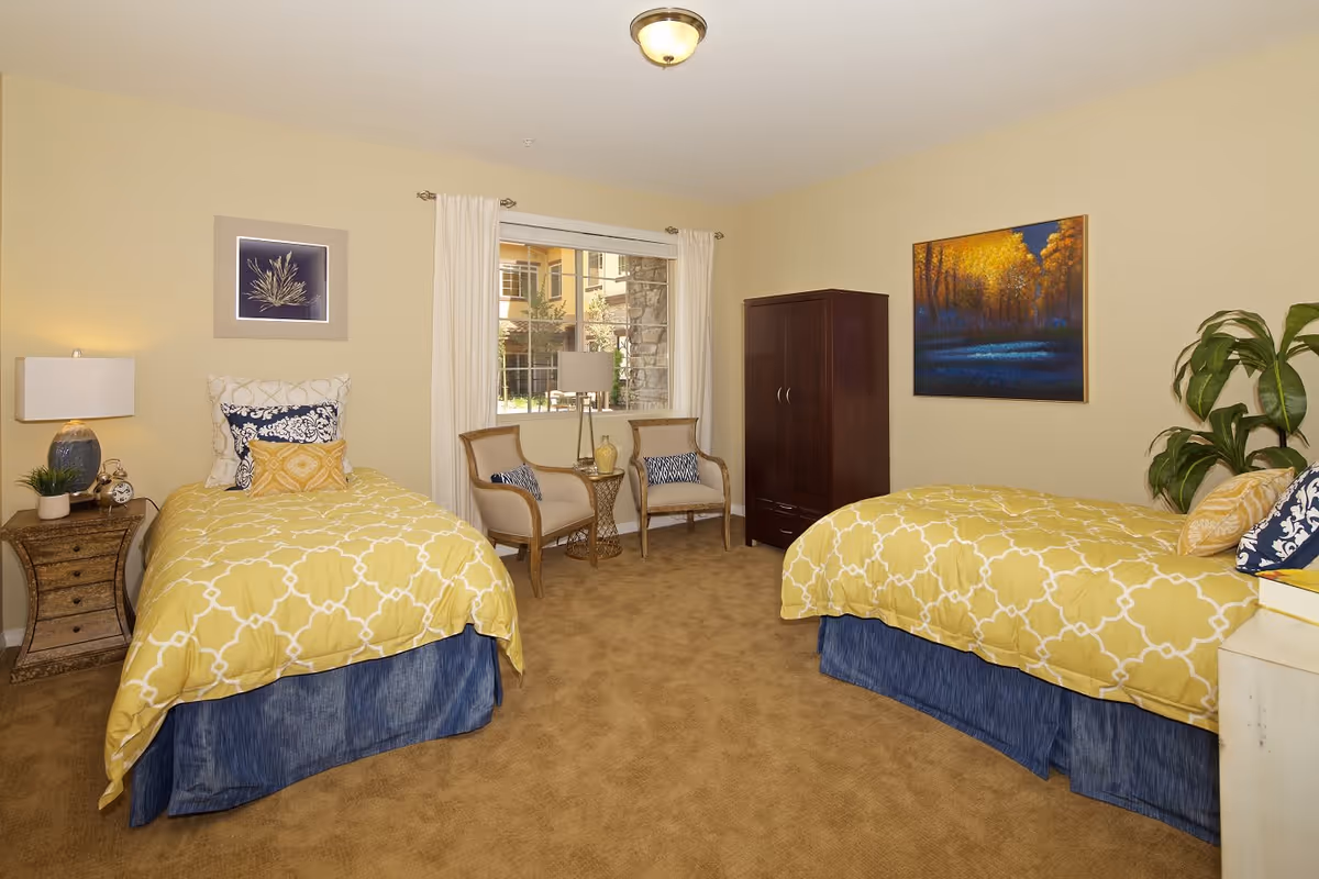A bedroom with two twin beds featuring yellow patterned bedspreads and blue bed skirts. Between the beds is a wooden nightstand with a lamp and a small plant. There are two chairs with cushions and a small round table in front of a window with white curtains. A dark wooden wardrobe stands against the wall, and a painting of a forest with yellow and blue tones hangs above one bed. The room has beige walls and carpeted floor.