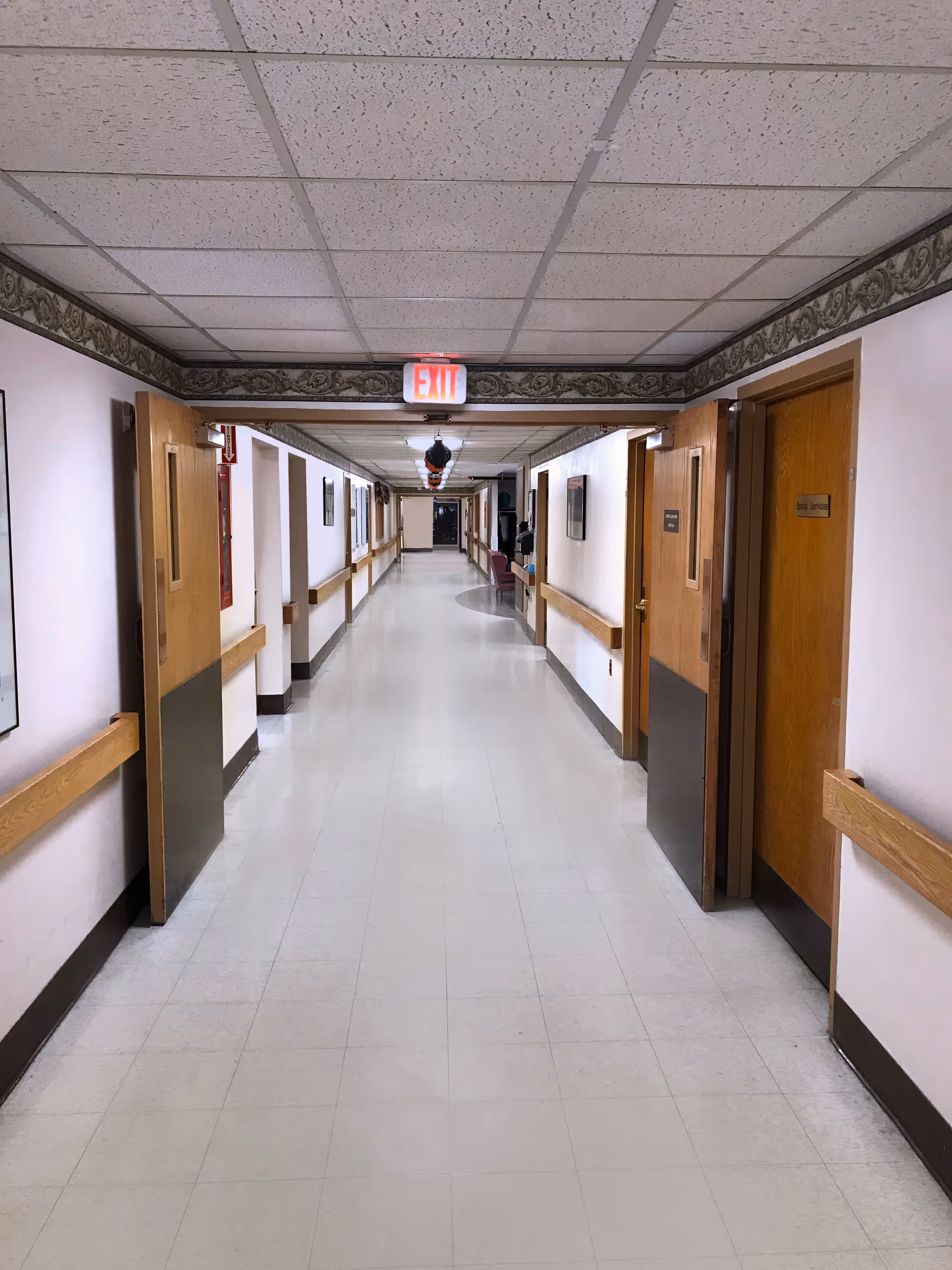 Empty institutional hallway with tiled ceiling, linoleum floor, wooden doors and handrails, and an EXIT sign above the distant doorway.