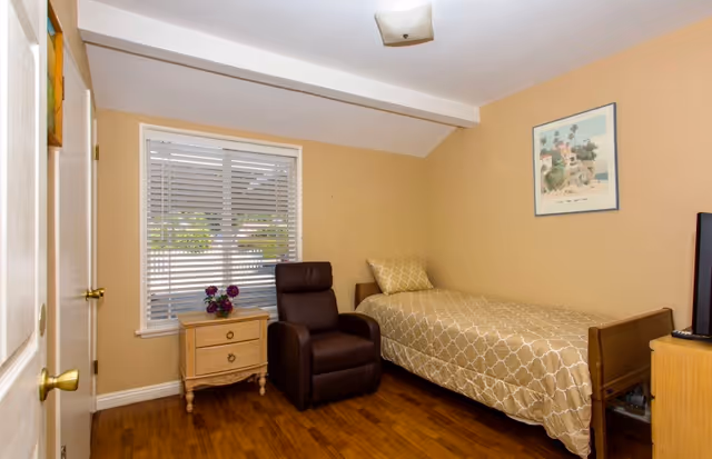 Small furnished bedroom with a single bed, recliner chair, nightstand, and window with blinds.