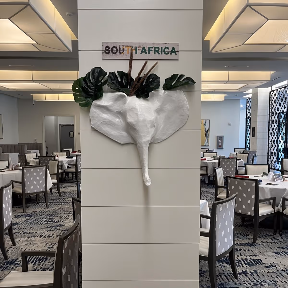 Interior view of a dining room with multiple tables and chairs arranged neatly. A white wall panel in the center features a decorative elephant head sculpture with green leaves and a sign above it that reads 'SOUTH AFRICA'. The room has modern lighting fixtures and patterned carpet flooring.
