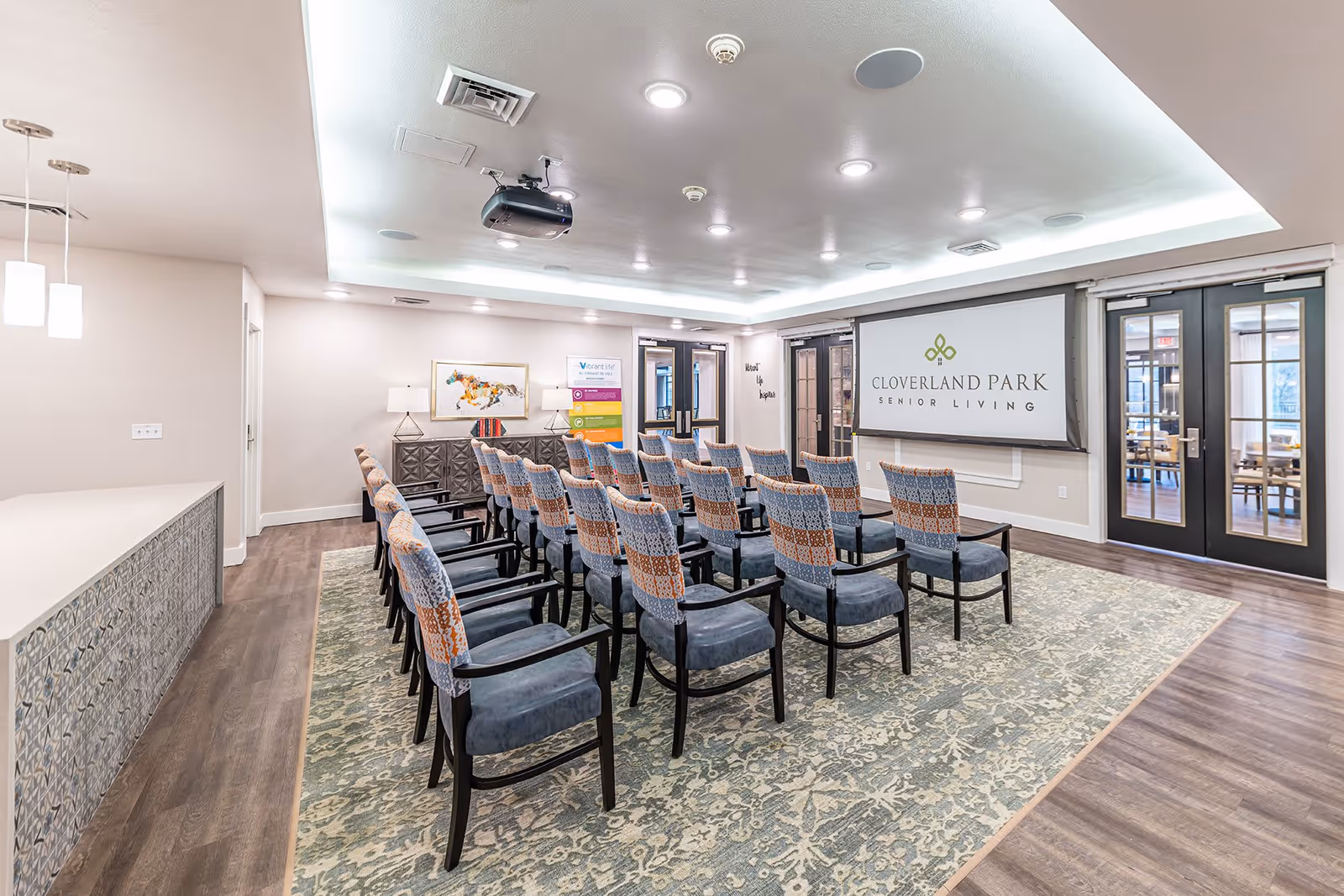 A well-lit meeting or presentation room with rows of patterned chairs facing a large screen displaying the Cloverland Park Senior Living logo. The room features a decorative rug, wooden flooring, a ceiling-mounted projector, and modern lighting. There are double glass doors and a sideboard with lamps and artwork on the wall.