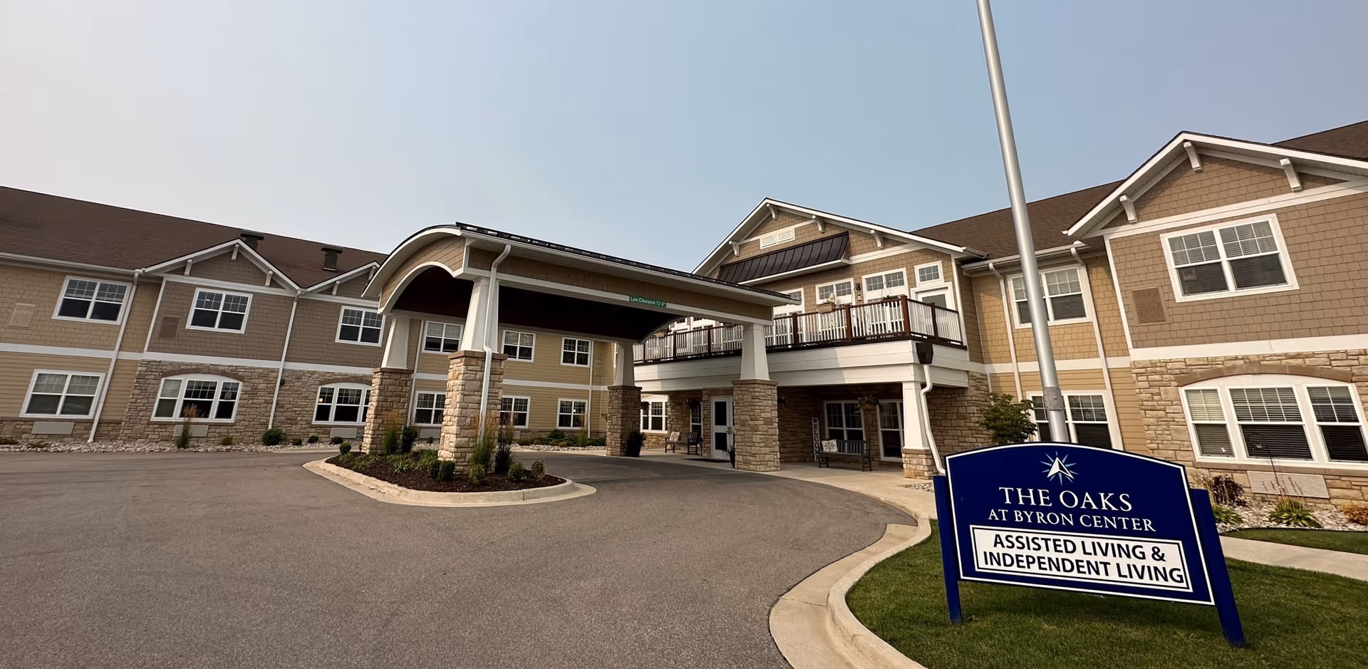 Exterior view of The Oaks at Byron Center, a senior living facility with a covered entrance supported by stone pillars, beige siding, white trim, and multiple windows. A blue sign in front reads 'The Oaks at Byron Center Assisted Living & Independent Living.'
