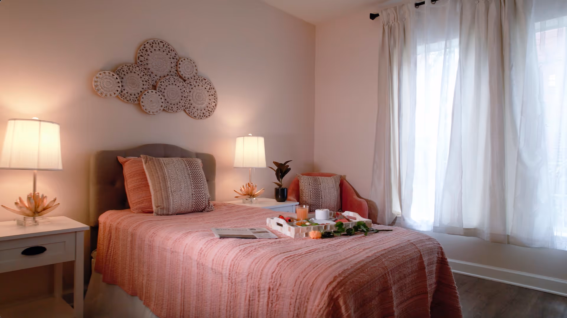 A cozy bedroom featuring a single bed with pink bedding, bedside tables with lamps, decorative wall art, and a curtained window.