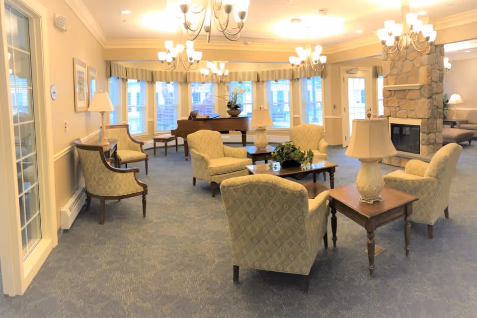 A bright and spacious living room area with multiple upholstered armchairs arranged around wooden coffee tables. The room features a stone fireplace, several table lamps, chandeliers hanging from the ceiling, large windows with valances, and a grand piano near the windows.