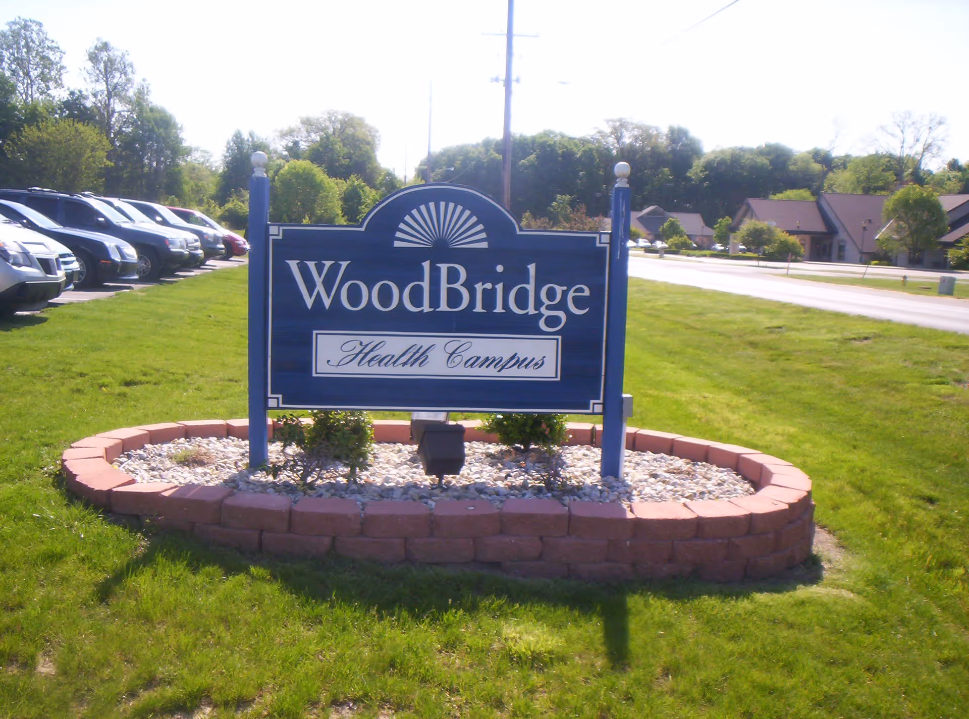 A blue sign with white text that reads 'WoodBridge Health Campus' situated on a grassy area bordered by a low brick wall with small plants and white rocks inside. There are parked cars to the left and buildings in the background under a clear sky.