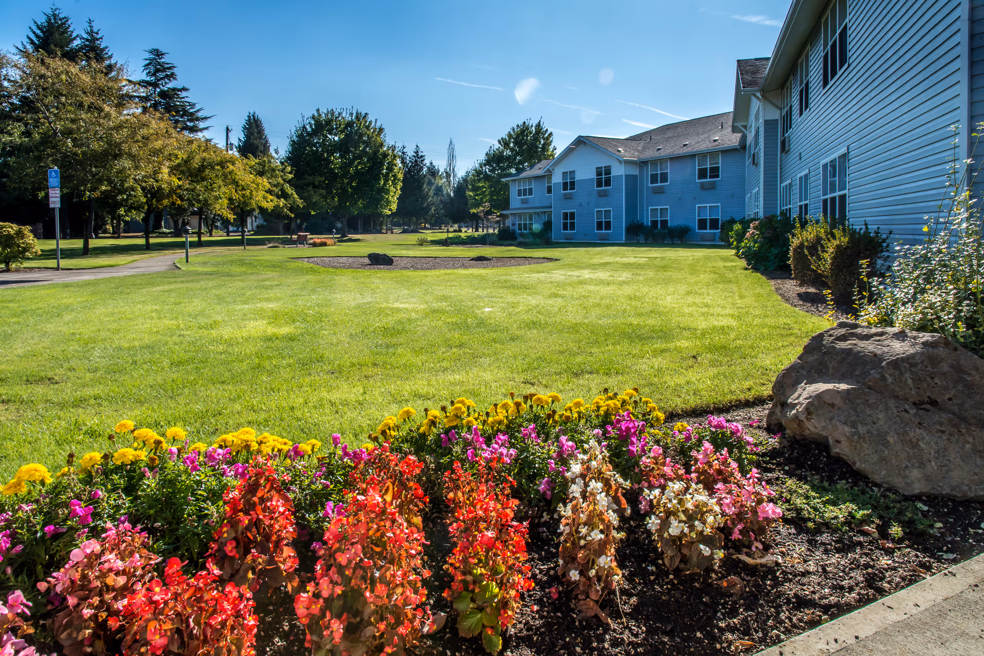 Colorful flower bed and green lawn in front of a multi-story assisted living building under a clear blue sky.