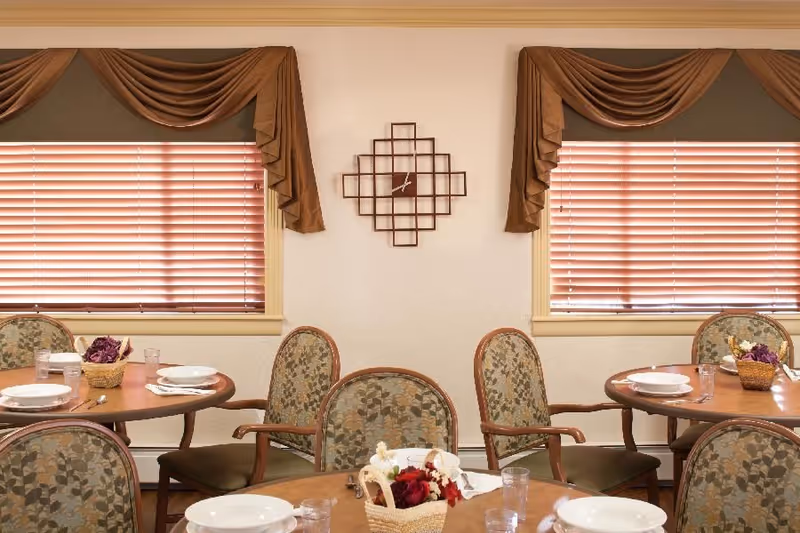 Communal dining room with round tables set with plates and glasses, patterned chairs, window blinds, and a decorative wall clock.