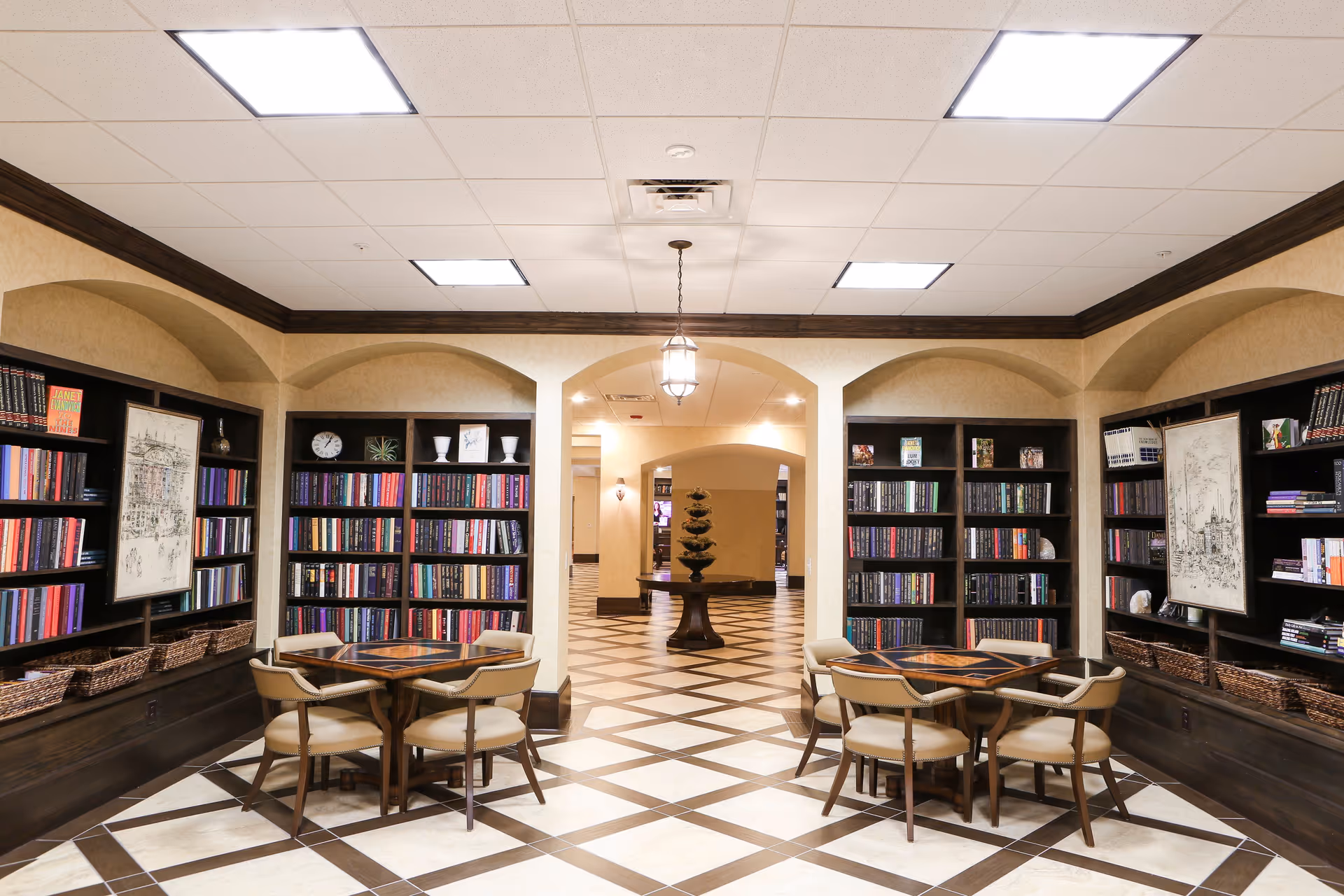 Interior view of a library or reading area in a senior living facility with bookshelves filled with books along the walls, two small tables each surrounded by four chairs, and a decorative table with a plant in the background. The floor has a checkered tile pattern and the ceiling has recessed lighting.
