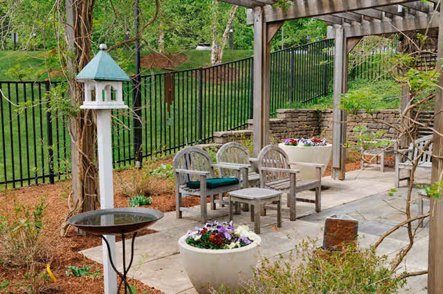 Outdoor patio area with wooden chairs and small tables under a pergola, surrounded by flower pots and greenery, with a birdhouse and birdbath nearby.