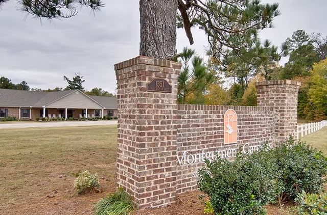 Brick entrance sign for Montgomery Gardens senior living facility with a large tree behind it, bushes in front, and a single-story building in the background surrounded by grass and trees.
