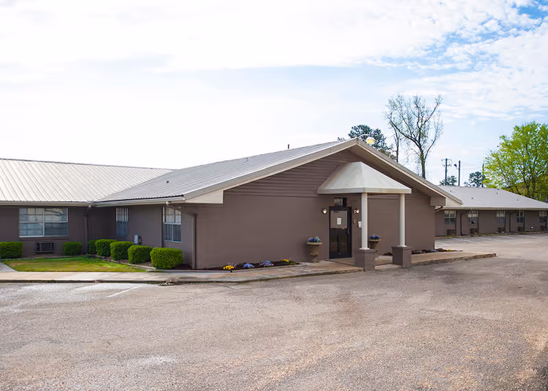 Exterior view of a single-story building with a sloped metal roof, brown brick walls, and a small covered entrance supported by two columns. There are small bushes and flower beds near the building, and a paved parking area in front. Trees and a partly cloudy sky are visible in the background.