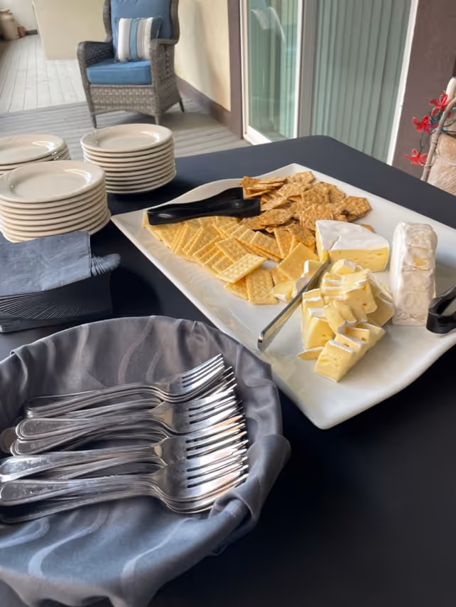 A table set with a platter of assorted crackers and cheeses, a stack of white plates, a basket lined with a gray cloth holding forks, and a blue cushioned wicker chair in the background on a porch or patio area.