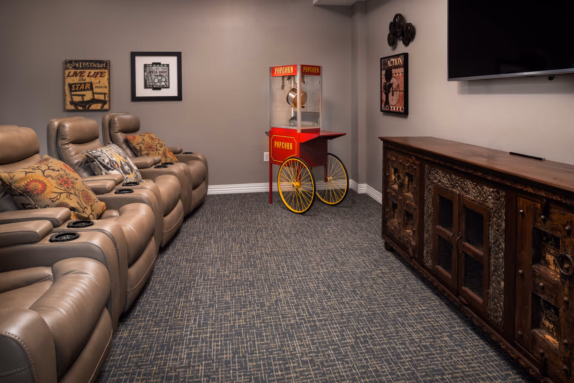 A cozy home theater room with four beige leather recliner chairs adorned with decorative pillows, a vintage-style red popcorn machine with yellow wheels in the corner, a large flat-screen TV mounted on the wall, and a rustic wooden cabinet beneath the TV. The walls are decorated with framed movie-themed artwork and film reels.