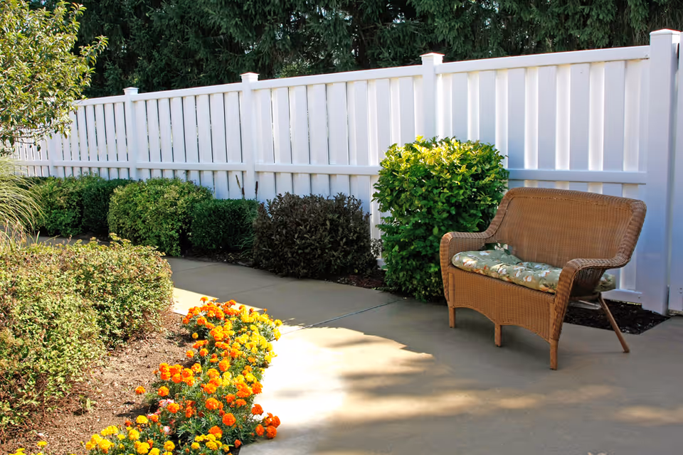 Outdoor garden area with a white fence, green bushes, and a flower bed with orange and yellow flowers. A wicker bench with a floral cushion is placed on a concrete pathway.