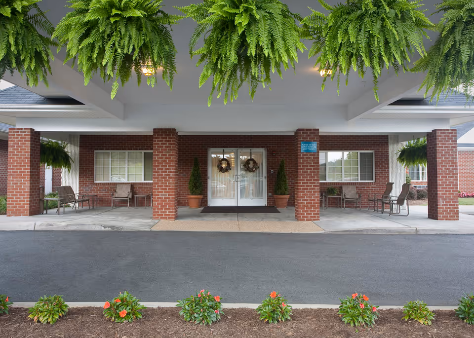Entrance of The Laurels of Charlottesville facility with a covered driveway supported by brick pillars, hanging green ferns, potted plants, and seating areas on both sides of the double glass doors decorated with wreaths.