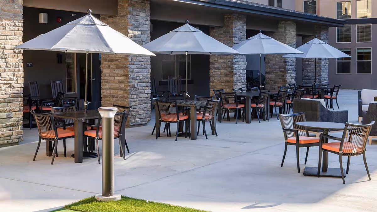 Outdoor patio area at Hilltop Springs Senior Living with multiple tables and chairs, each table shaded by a large white umbrella. The patio is adjacent to a building with stone pillars and windows.