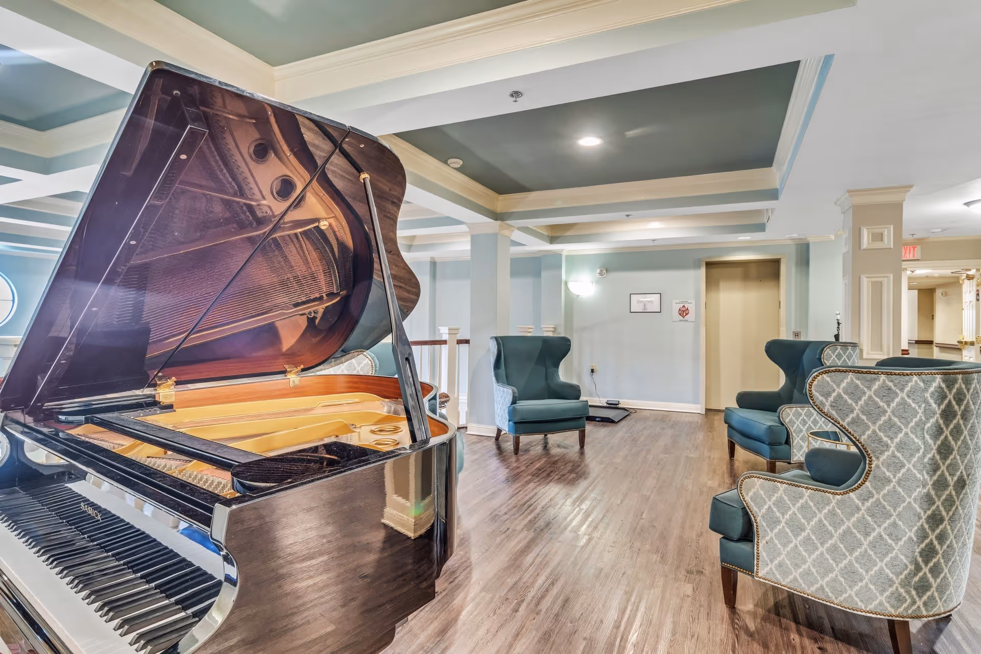 A bright and spacious common area with a grand piano in the foreground and several upholstered armchairs arranged around the room. The space features light blue walls, white trim, a coffered ceiling, and wood flooring. An elevator door is visible in the background.