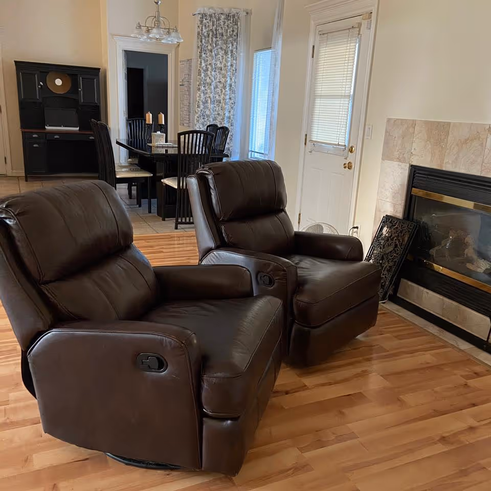 Two brown leather recliners on hardwood floors next to a fireplace with a dining area visible in the background.