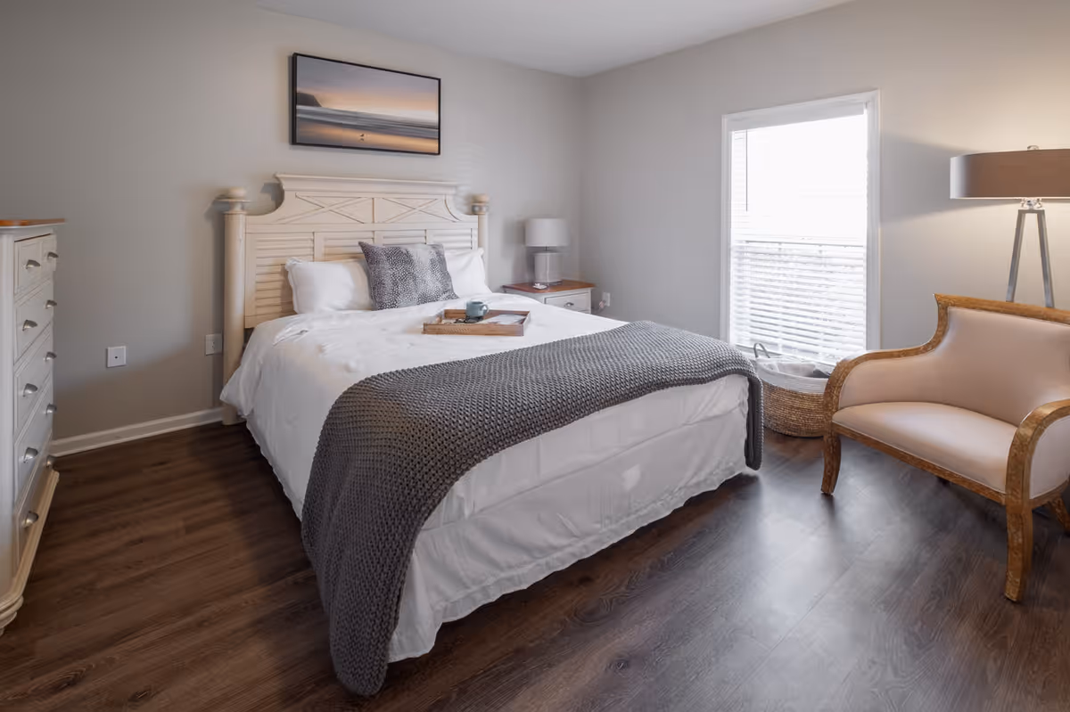 A neatly made bed with white bedding and a gray knitted throw blanket in a bedroom. There are two pillows on the bed, a wooden headboard, a nightstand with a lamp, a dresser, a window with blinds, a basket, and a cushioned armchair with wooden frame. The floor is dark wood and the walls are light gray.