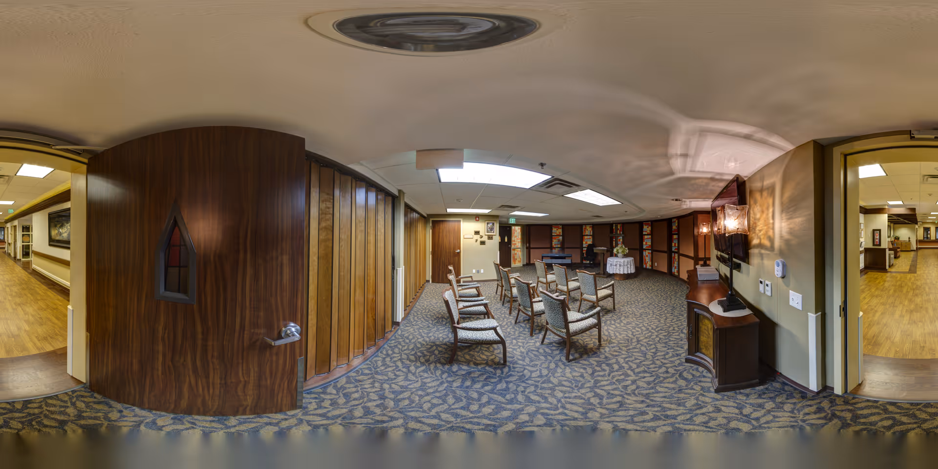 Interior view of a meeting or activity room in a senior living facility. The room has patterned carpet flooring and several wooden chairs with cushions arranged in rows facing a wall-mounted TV. There is a small table with a flower arrangement and a piano against the far wall. The room is well-lit with ceiling lights, and there are two open doorways leading to hallways with wood flooring.