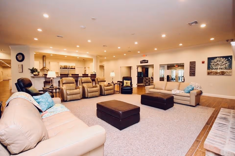 A spacious and well-lit living room area in an assisted living facility featuring beige leather sofas, multiple recliner chairs, two brown ottomans, a carpeted floor, and a kitchen area with bar stools in the background. The walls are decorated with artwork and a welcome sign is visible above a doorway.
