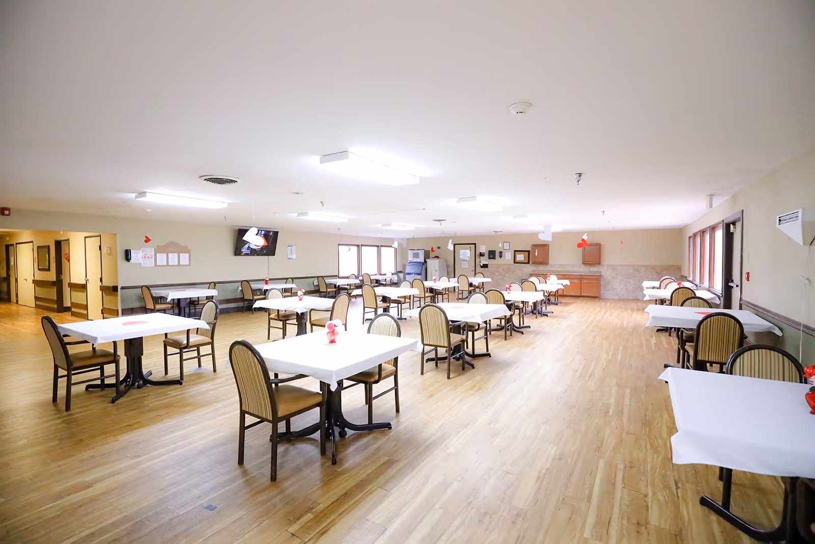 A spacious dining room with multiple tables covered in white tablecloths, each surrounded by chairs. The room has wooden flooring, large windows on the right side, and overhead fluorescent lighting. There is a television mounted on the far wall and a kitchenette area with cabinets and a refrigerator in the back.