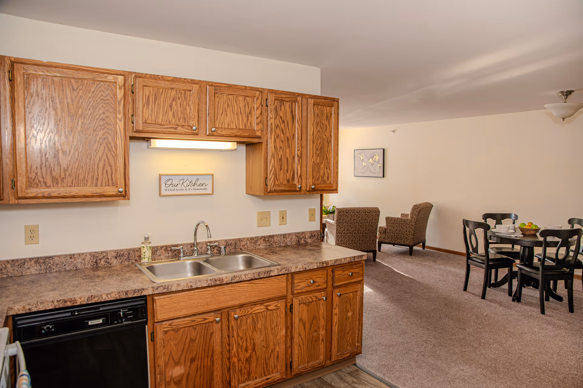 Interior view of a kitchen area with wooden cabinets, a double sink, and a countertop. In the background, there is a dining area with a round table and four chairs, and a seating area with two upholstered armchairs. The walls are light-colored and there is a framed artwork on the wall.