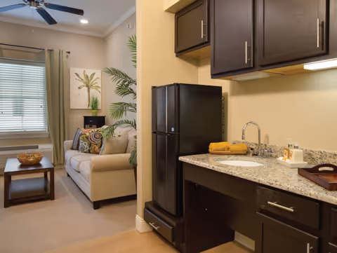 Cozy studio interior with a small kitchenette featuring granite countertops, a mini fridge and sink, opening to a living area with a sofa and coffee table.