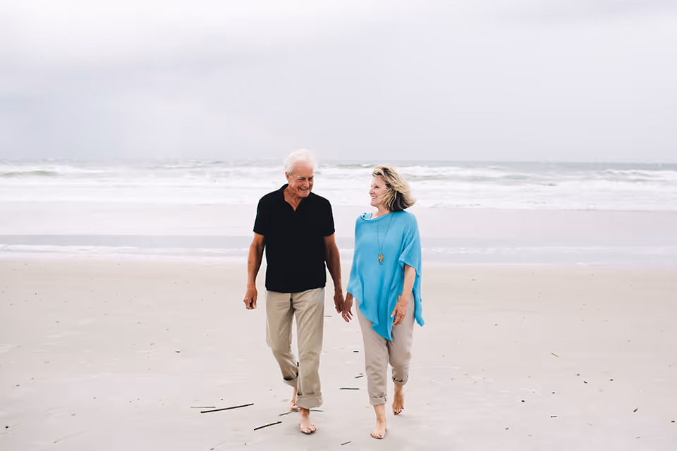 An elderly couple walking barefoot on a sandy beach near the ocean, holding hands and smiling at each other under a cloudy sky.