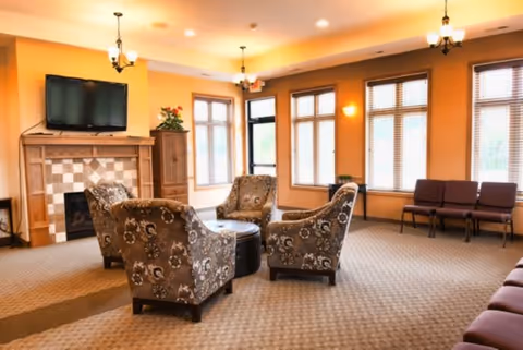 A cozy living room area with four patterned armchairs arranged around a round ottoman in front of a fireplace with a mounted flat-screen TV above it. The room has large windows with blinds, warm yellow walls, carpeted floor, and ceiling lights with chandeliers.