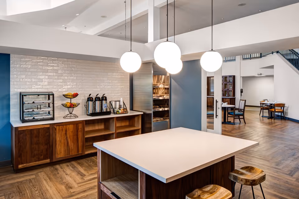 A modern dining area in Holiday Rock Creek featuring a central island with two wooden stools, a countertop with coffee dispensers, fruit baskets, and a display case. The space has wooden flooring, white walls with blue accents, and three round pendant lights hanging from the ceiling. In the background, there are tables and chairs arranged for dining.