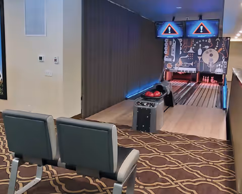Indoor bowling alley with two lanes, bowling balls on a rack, and two screens displaying warning signs above the pins. Two gray chairs are positioned in front of the lanes on a patterned carpeted floor.
