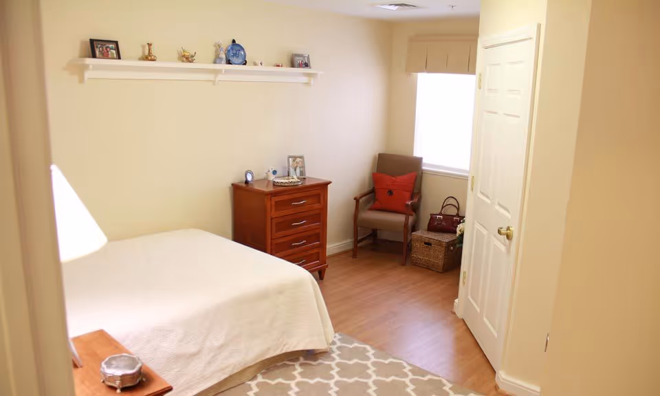 A cozy bedroom with a bed covered in a white quilt, a wooden nightstand with a lamp, a wooden dresser with decorative items on top, a chair with a red pillow, a basket, and a window letting in natural light. A white door is visible on the right side of the room.
