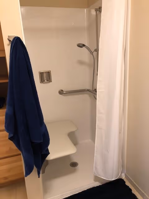 A small shower area with a white built-in seat, a handheld showerhead attached to a vertical bar, a grab bar on the wall, a soap holder, and a white shower curtain. A blue towel hangs on a hook outside the shower, and a black bath mat is on the floor.