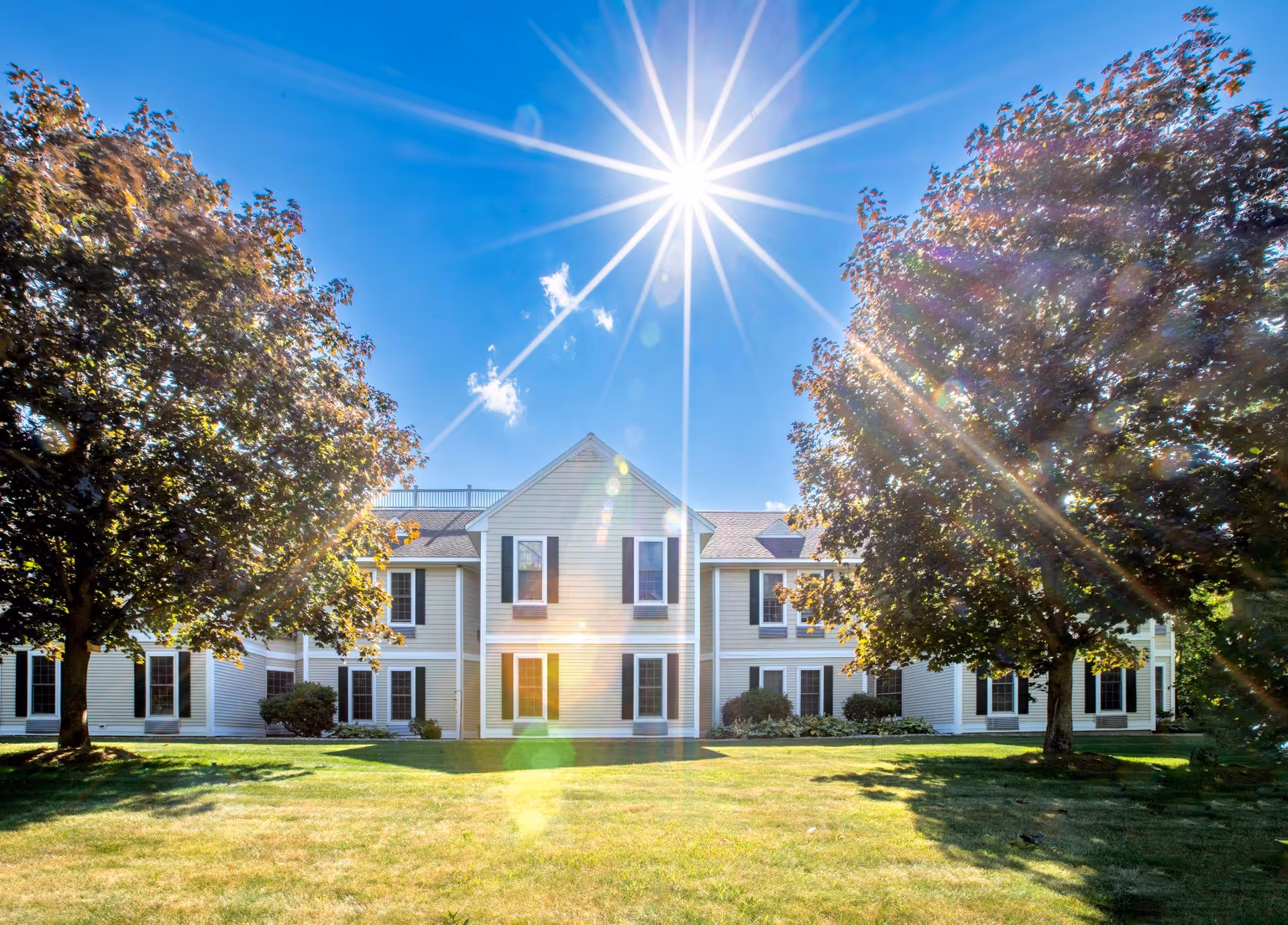 Exterior view of a two-story residential building with white siding and black shutters, surrounded by green grass and large trees under a bright sun in a clear blue sky.
