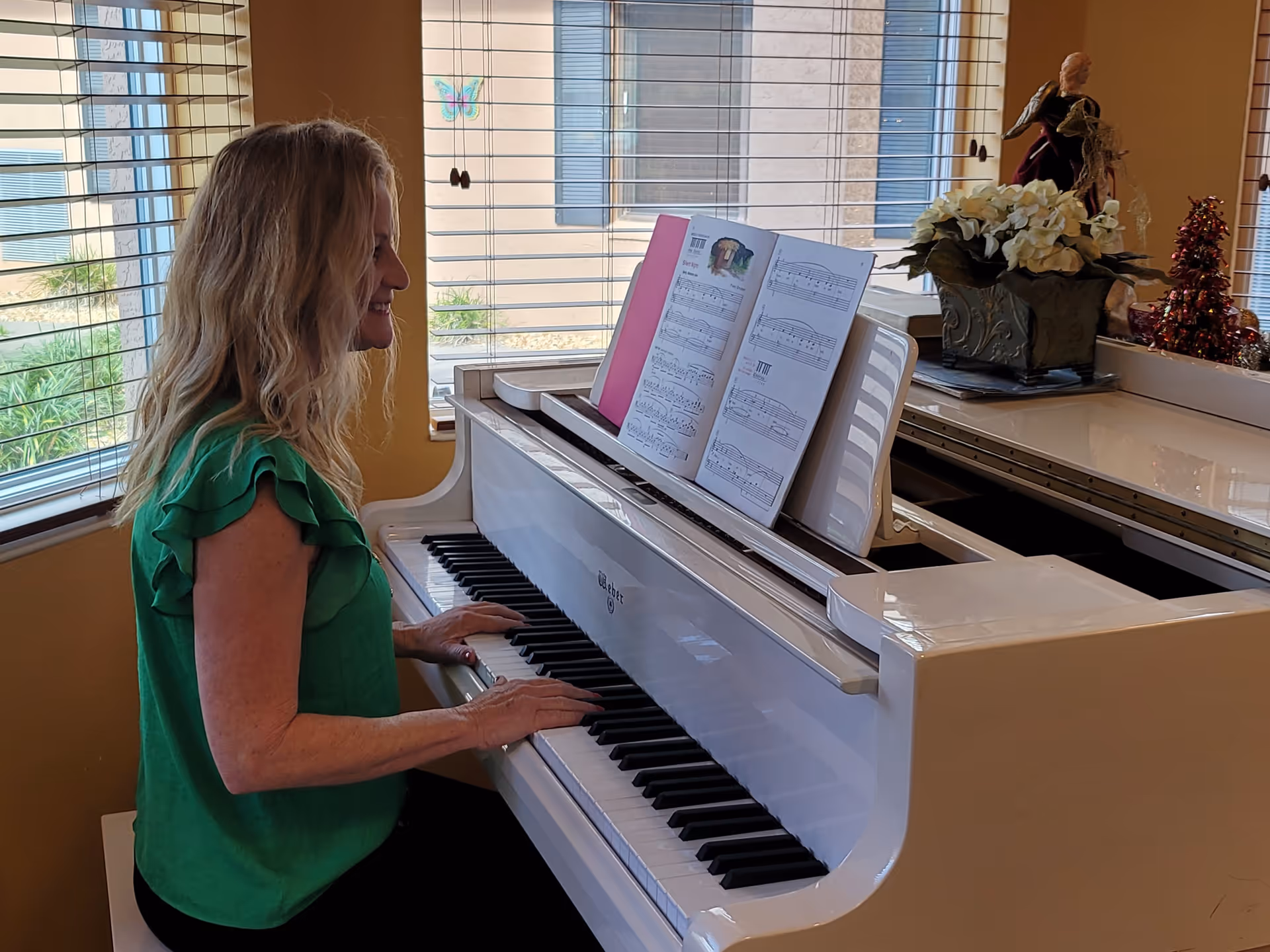 A woman with blonde hair wearing a green blouse is sitting and playing a white grand piano in a room with large windows and beige walls. There is sheet music on the piano and decorative items, including a flower arrangement and figurines, on top of the piano.