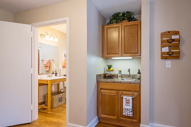 Small kitchenette area with wooden cabinets, a countertop with a sink, soap dispenser, and a two-tier fruit basket. To the left, an open door reveals a bathroom with a wooden vanity, mirror, and light fixtures above it. A wicker basket is visible under the bathroom vanity.